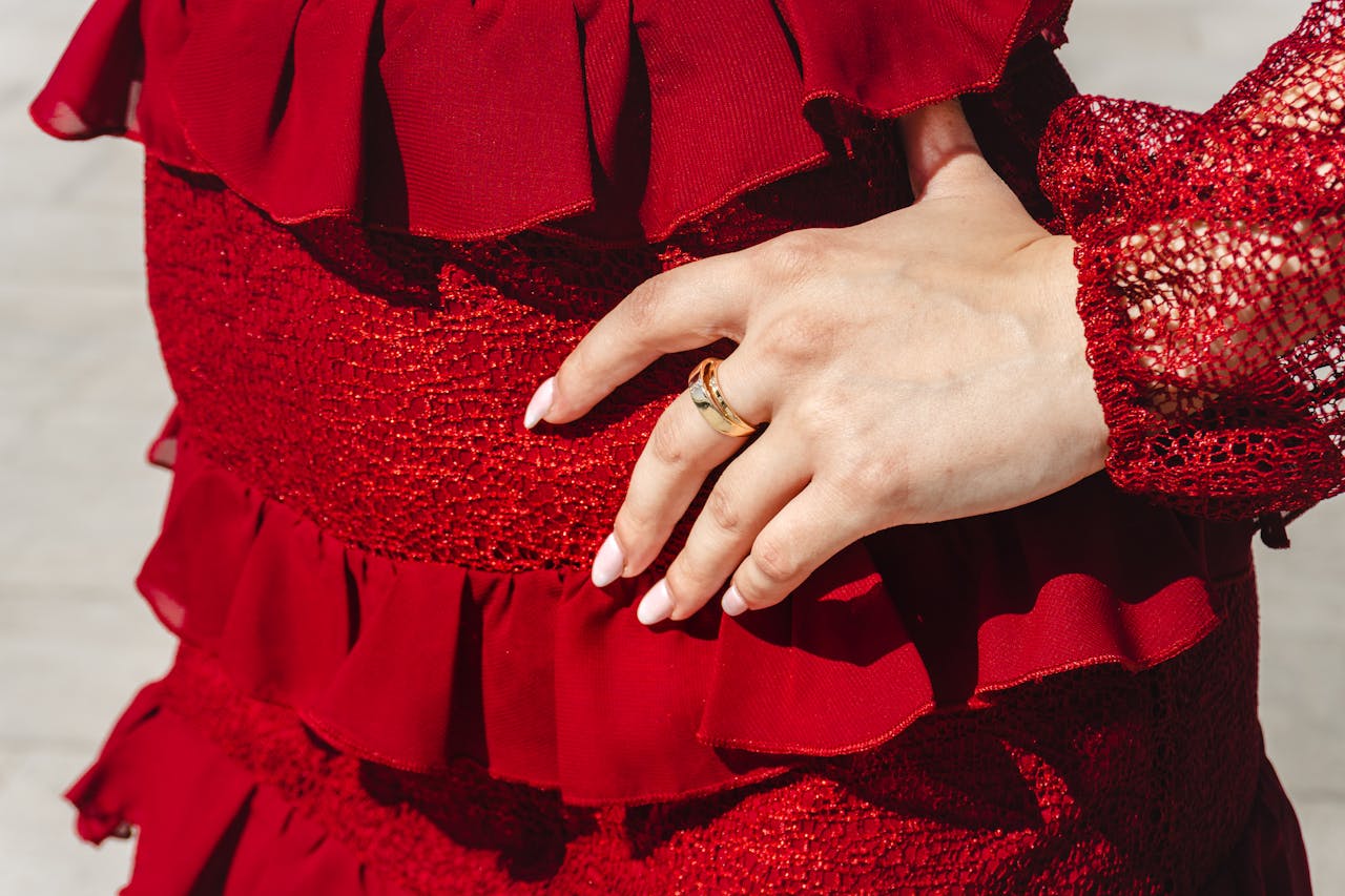 Home Detailed view of a hand with ring resting on a textured red dress, showcasing elegance and style.