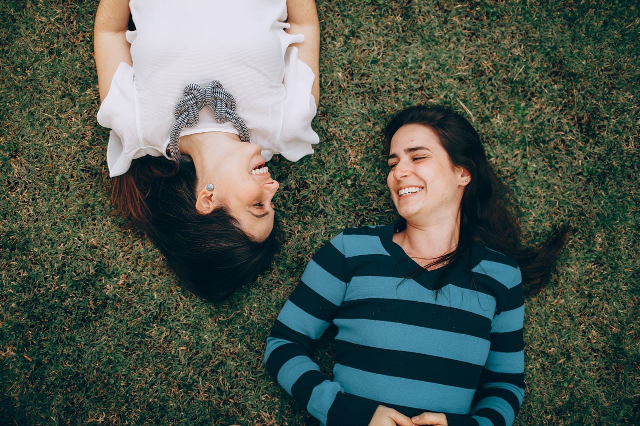 About Two women lie on the grass, sharing laughter and happiness in a candid outdoor moment.